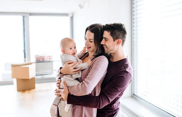 A portrait of young couple with a baby and cardboard boxes moving in a new home. — Stock Photo, Image
