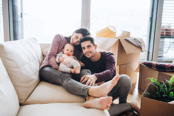 A portrait of young couple with a baby and cardboard boxes moving in a new home. — Stock Photo, Image