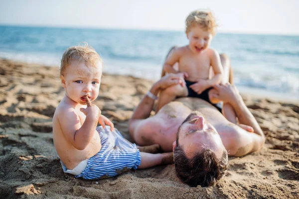 Un padre con due bambini piccoli sdraiati sulla spiaggia di sabbia in vacanza estiva . — Foto Stock