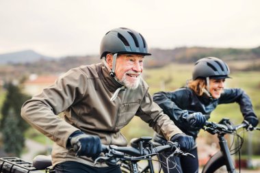 Active senior couple with electrobikes cycling outdoors on a road in nature. clipart