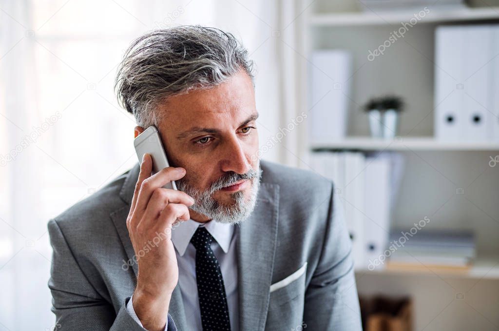 Serious mature businessman with smartphone sitting at the table, making a phone call.