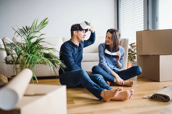 Una joven pareja con gafas VR sentada en el suelo, mudándose a un nuevo hogar . — Foto de Stock