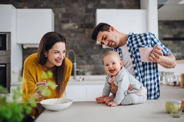 Una giovane famiglia a casa, un uomo che tiene in braccio un bambino e una donna che le dà da mangiare . — Foto Stock
