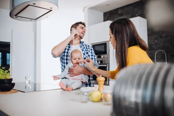 スプーンであげて女と赤ん坊を保持しているスマート フォンを持つ男 自宅の台所で立っている若い家族の肖像画 — ストック写真
