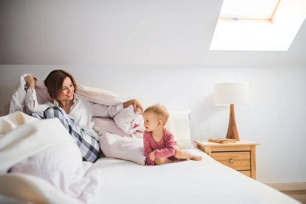 En ung mor med lilla dotter sitter inomhus på sängen på morgonen, spela. — Stockfoto