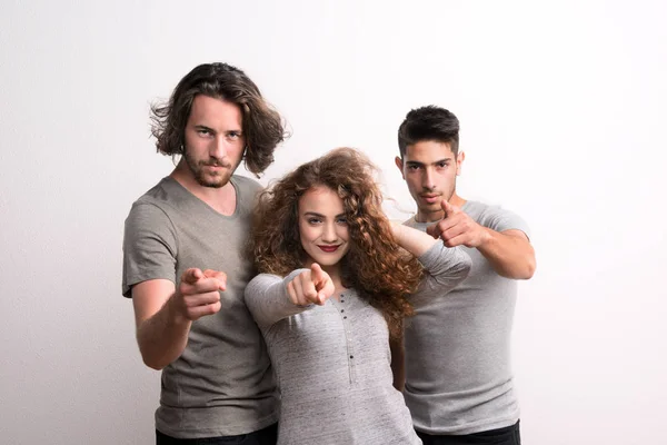 Retrato de una joven alegre con dos amigos varones de pie en un estudio, señalándote . — Foto de Stock