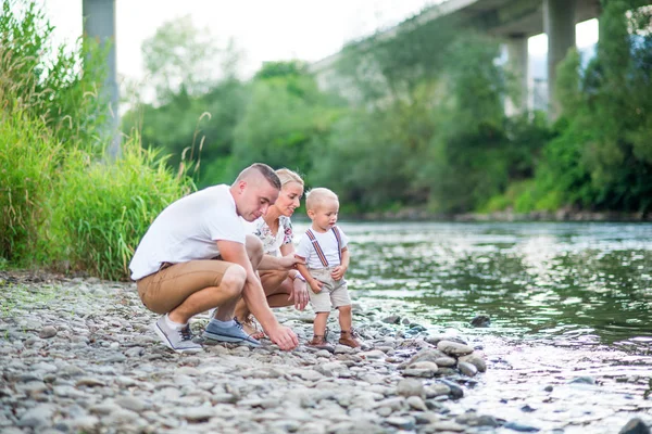 Família jovem com um menino pequeno na natureza ensolarada do verão, jogando pelo rio . — Fotografia de Stock