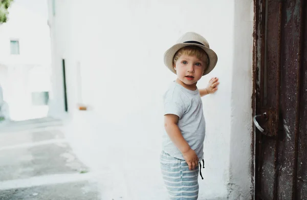 Un piccolo bambino con cappello in piedi accanto a una casa in città in vacanza estiva . — Foto Stock