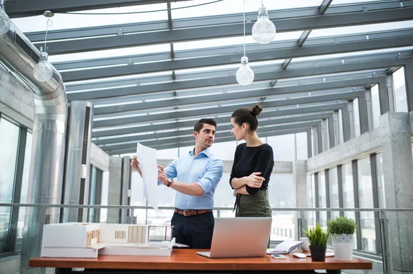 Dos jóvenes arquitectos con planos y modelo de una casa de pie en la oficina, hablando . — Foto de Stock