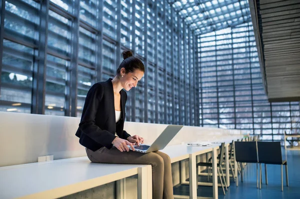 Joven estudiante o mujer de negocios sentada en el escritorio en la habitación de una biblioteca u oficina, usando una computadora portátil . — Foto de Stock