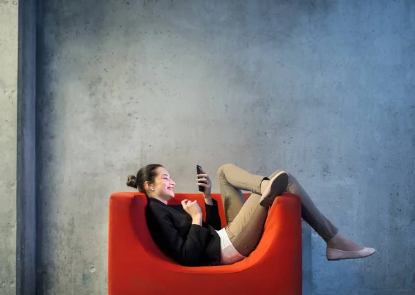 En ung affärskvinna med smartphone som sitter på röd fåtölj i office. — Stockfoto