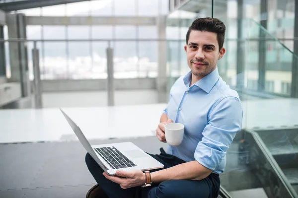 Um retrato de jovem empresário com laptop sentado no corredor fora do escritório . — Fotografia de Stock