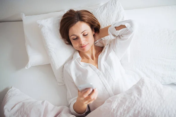 Een bovenaanzicht van jonge vrouw liggend in bed binnenshuis in een slaapkamer, met behulp van smartphone. — Stockfoto