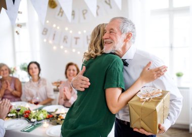 Young woman giving a gift to her grandfather on indoor party, hugging. clipart