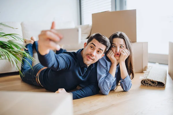 A young couple with a smartphone moving in a new home, taking selfie and making a face. — Stock Photo, Image