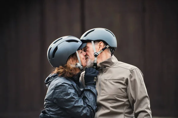 Ativo casal sênior com capacetes de bicicleta em pé ao ar livre againts fundo escuro, beijando . — Fotografia de Stock