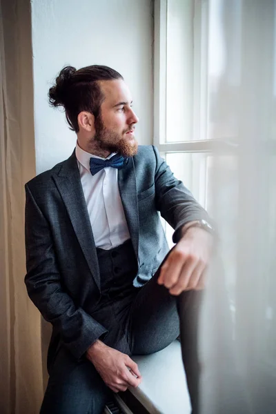 A handsome hipster young man with formal suit sitting on a window sill on an indoor party. — Stock Photo, Image