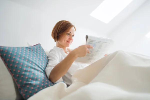 朝の寝室のベッドで新聞を読む若い女性. — ストック写真