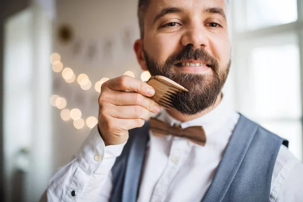 Hipster homem em pé dentro de casa em um conjunto de quarto para uma festa, pentear a barba . — Fotografia de Stock