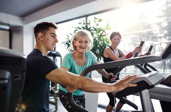 Dos alegres mujeres mayores en el gimnasio con un joven entrenador haciendo ejercicio cardiovascular . — Foto de Stock