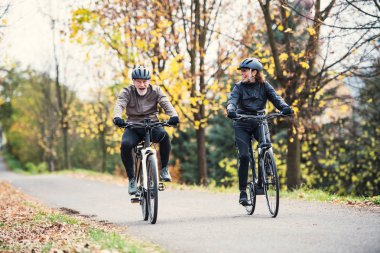 A senior couple with electrobikes cycling outdoors on a road in park in autumn. clipart