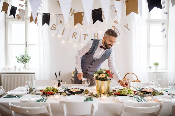 Portrait d'un homme mûr à l'intérieur dans une pièce aménagée pour une fête, mettant la table . — Photo