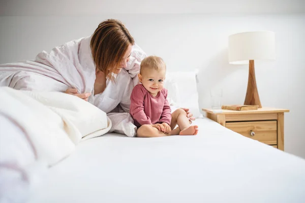 Молодая мать с маленькой дочерью сидит в помещении на кровати по утрам, играет . — стоковое фото