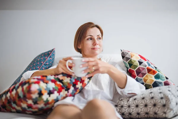 Молодая женщина в ночной рубашке сидит в помещении на кровати по утрам, держа чашку . — стоковое фото