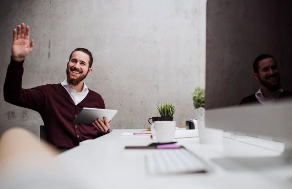 Un joven hombre de negocios con una tableta en la oficina, saludando a alguien . — Foto de Stock