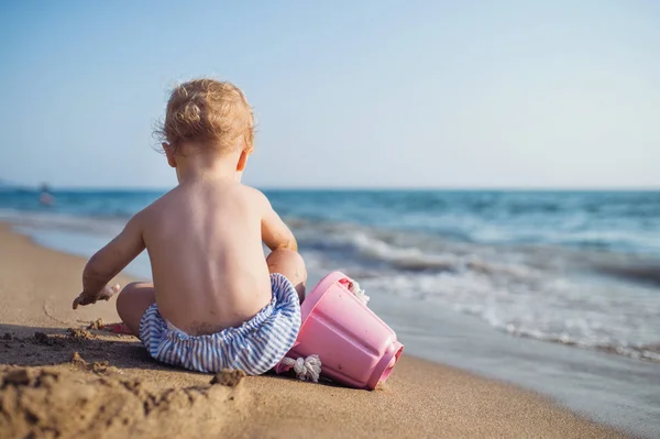 Una vista trasera de una niña pequeña sentada en la playa en las vacaciones de verano, jugando . —  Fotos de Stock