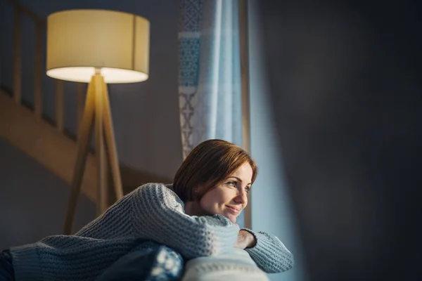 A young woman sitting indoors on a sofa at home, looking out of a window. — Stock Photo, Image