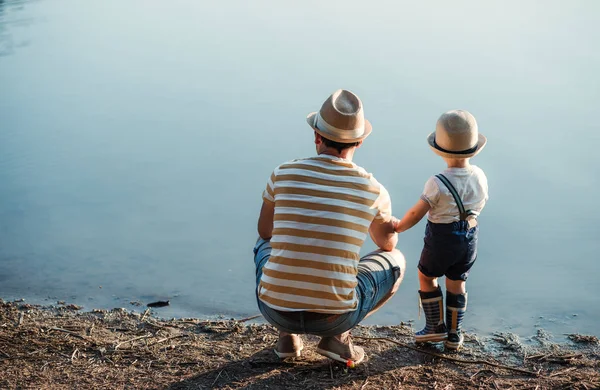 Una vista trasera del padre con un pequeño hijo pequeño que pasa tiempo al aire libre junto a un lago . —  Fotos de Stock