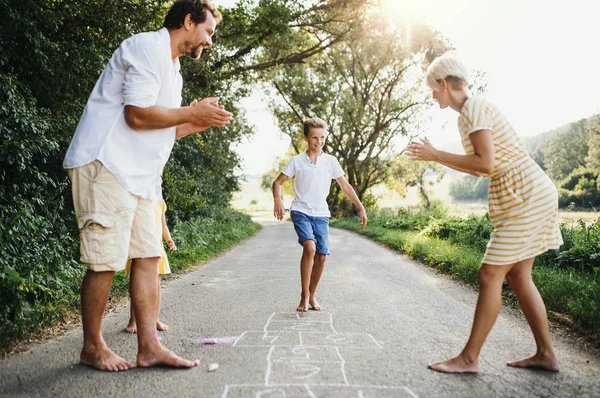 Une jeune famille avec de jeunes enfants jouant à la marelle sur une route en été . — Photo