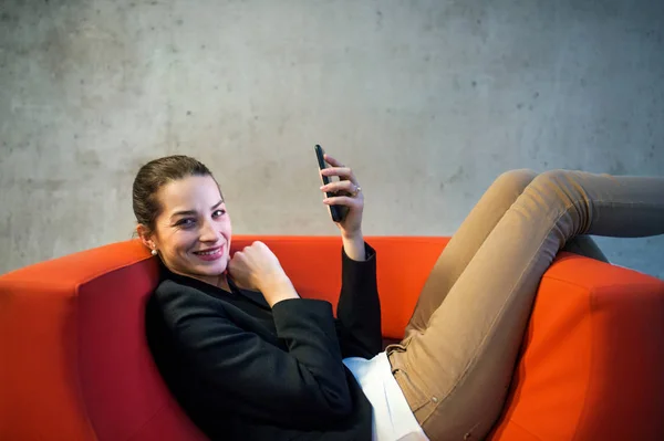 Seorang pengusaha muda dengan smartphone duduk di kursi merah di kantor . — Stok Foto