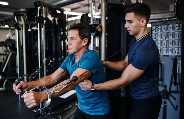 En äldre man med en ung tränare som gör styrka träning träning i gym. — Stockfoto