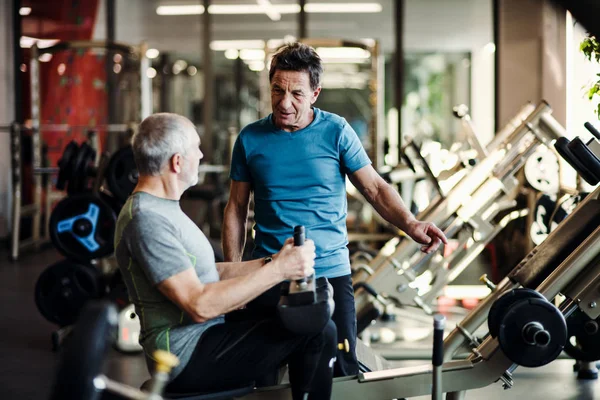 En äldre man med en ung tränare som gör styrka träning träning i gym. — Stockfoto