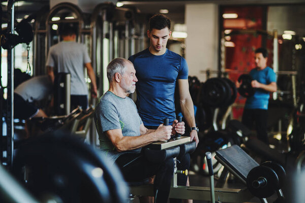 A senior man with a young trainer doing strength workout exercise in gym.