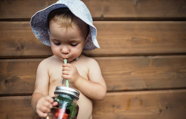 Ein kleines Mädchen mit einem Drink, das im Sommer vor einem hölzernen Hintergrund auf einer Terrasse steht. — Stockfoto