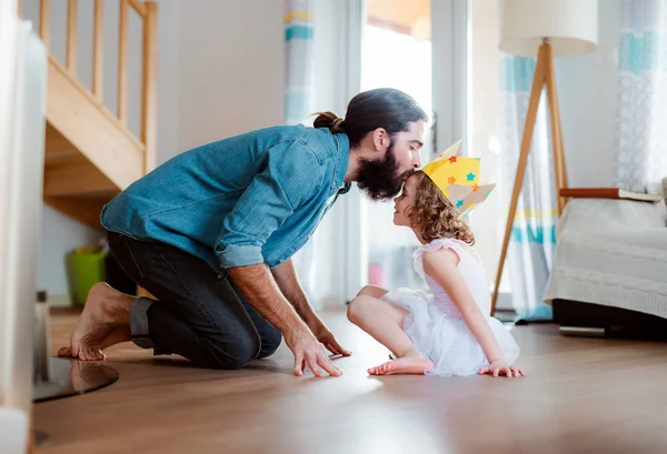 Una vista lateral de una niña pequeña con una corona de princesa y un padre joven en casa, jugando . — Foto de Stock