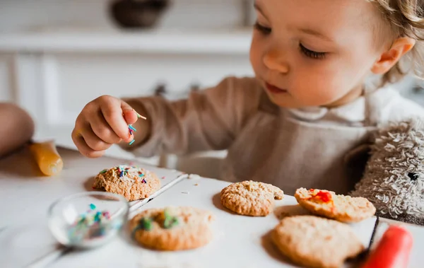 A small toddler girl sitting at the table, decorating cakes at home. — Stock Photo, Image