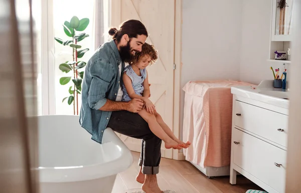 Uma menina pequena com o pai jovem no banheiro em casa, olhando para unhas pintadas . — Fotografia de Stock