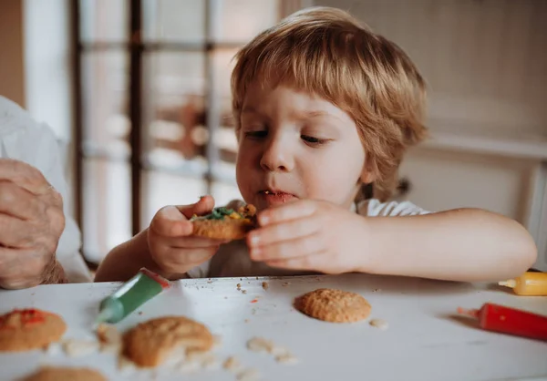 Un niño pequeño sentado a la mesa, decorando y comiendo pasteles en casa . —  Fotos de Stock