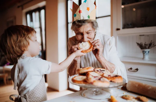 Starší babička s malým batoletem chlapce dělat a jíst dorty doma. — Stock fotografie