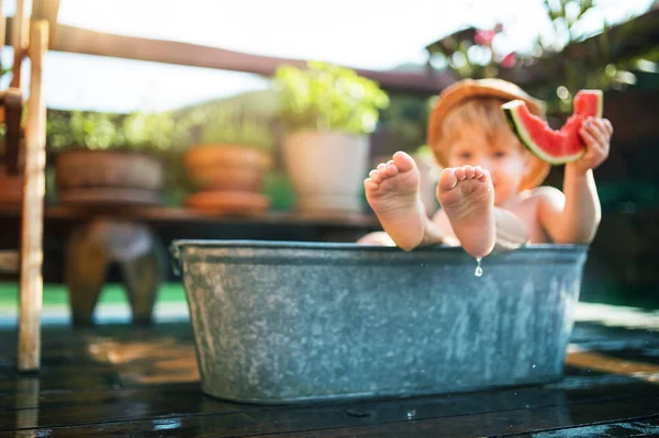 Mały chłopiec z czapką w kąpieli na świeżym powietrzu w ogrodzie w letni, jedzenie arbuza. — Zdjęcie stockowe