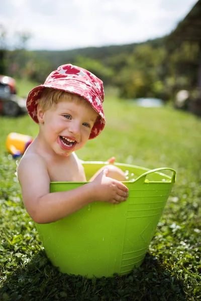 Small boy with a hat in bucket outdoors in garden in summer, playing in water. — Stock Photo, Image