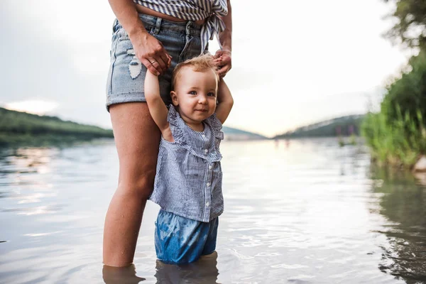 Una sección media de la madre con una hija pequeña al aire libre junto al río en verano . — Foto de Stock