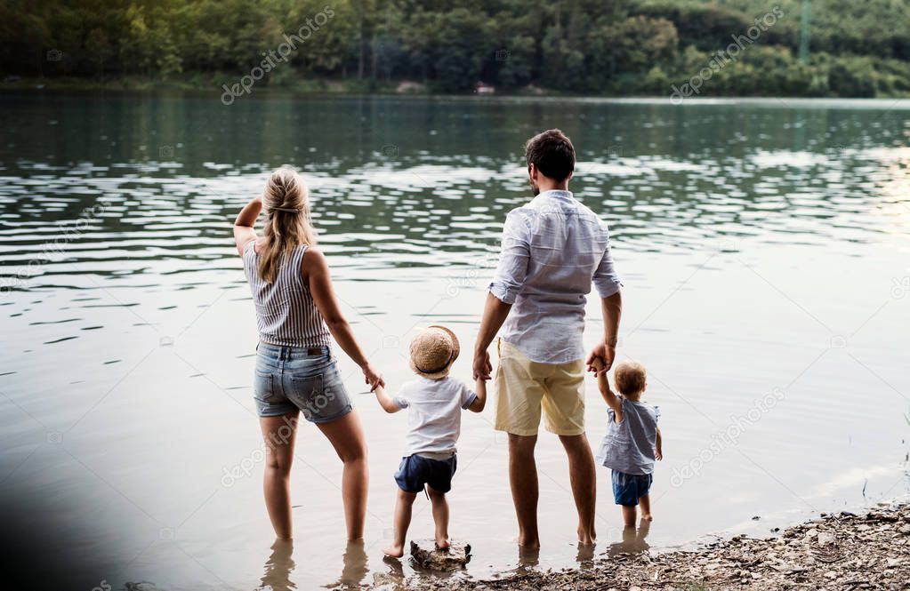 A rear view of family with two toddler children outdoors by the river in summer.