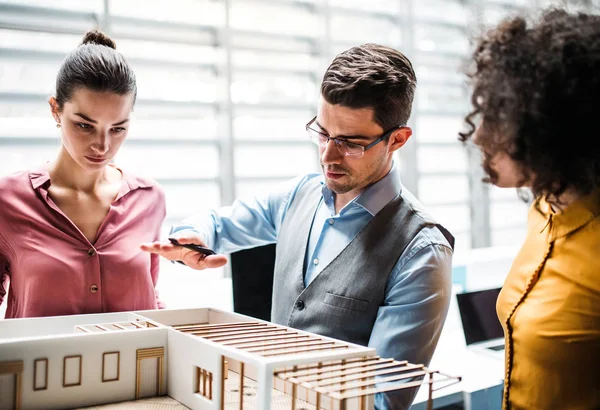 Group of young architects with model of a house standing in office, talking. — Stock Photo, Image