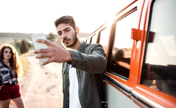 Um jovem casal com smartphone de carro em uma viagem pelo campo, tirando selfie . — Fotografia de Stock