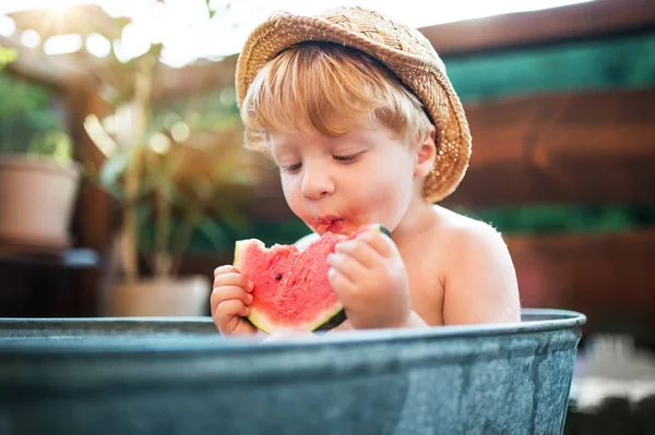 夏には、庭園で屋外のお風呂で帽子と小さな男の子はスイカを食べる. — ストック写真